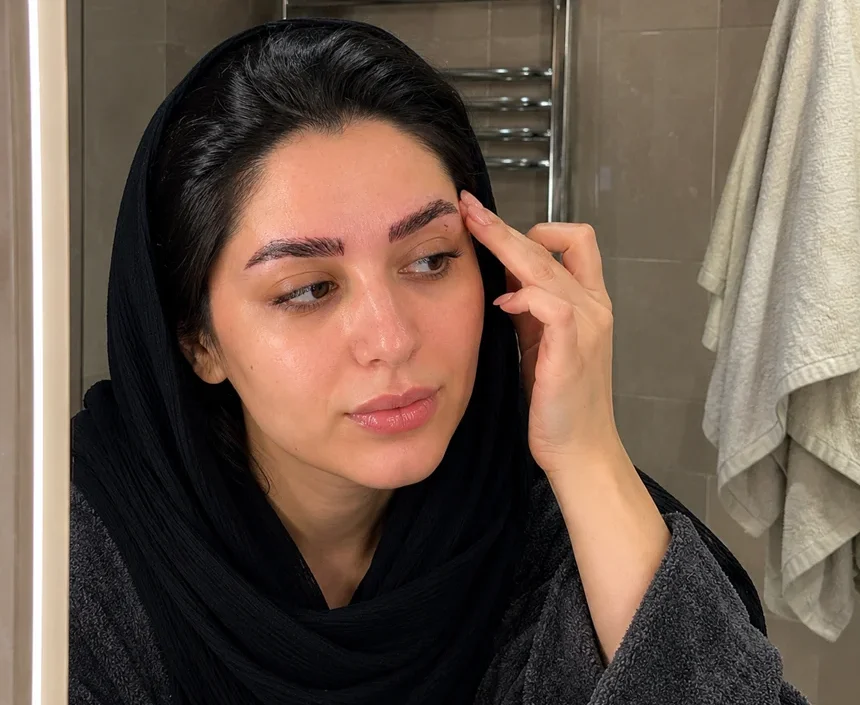 خانم ایرانی با روسری مشکی که به‌تازگی کاشت ابرو انجام داده، در مقابل آینه حمام در حال بررسی وضعیت ابروهای خود و کنترل علائم احتمالی عفونت یا التهاب پس از کاشت ابرو است.