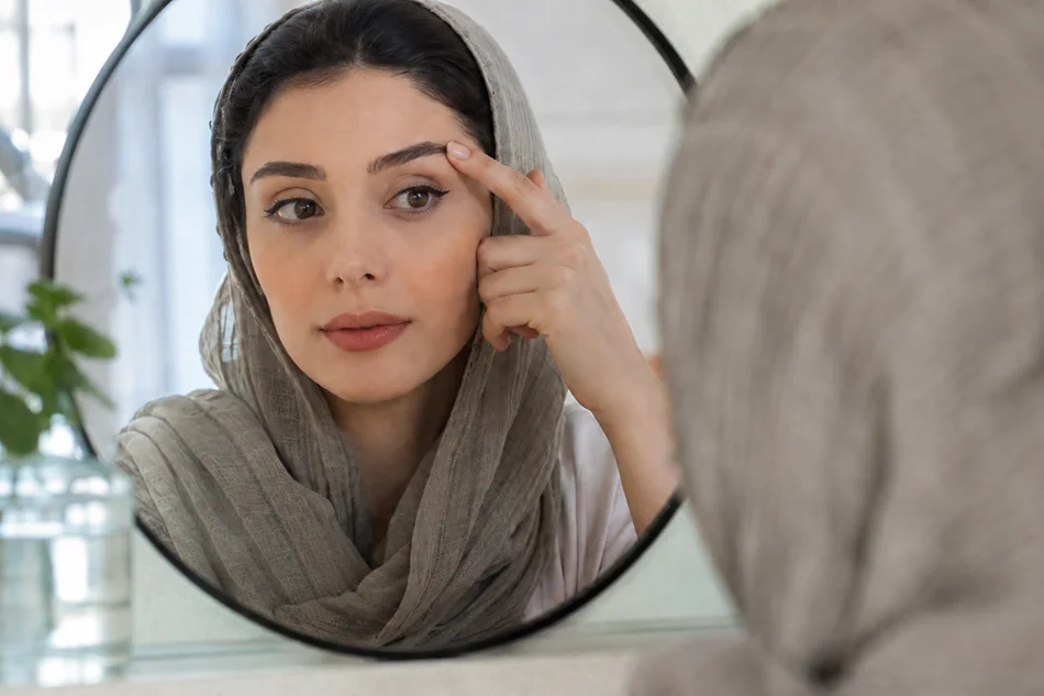 یک خانم با شال که به آینه نگاه میکنه و داره ابروهاشو چک میکنه